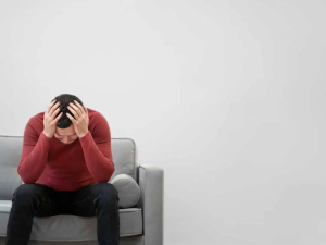 Feeling overwhelmed or anxious, a young man sits on a gray couch with his hands on his head, demonstrating stress relief and mental health support offered by Davenport Psychology.